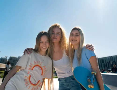 Three smiling teenage girls standing together outdoors, one holding a skateboard, symbolizing supportive friendships and peer connections in preventing substance use.